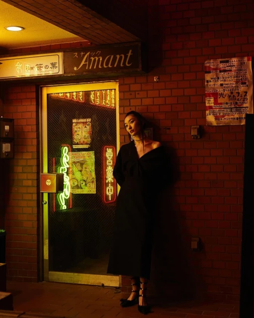 Adult female with dark hair poses in a black dress against a brick wall next to a doorway with neon lights in an urban setting.