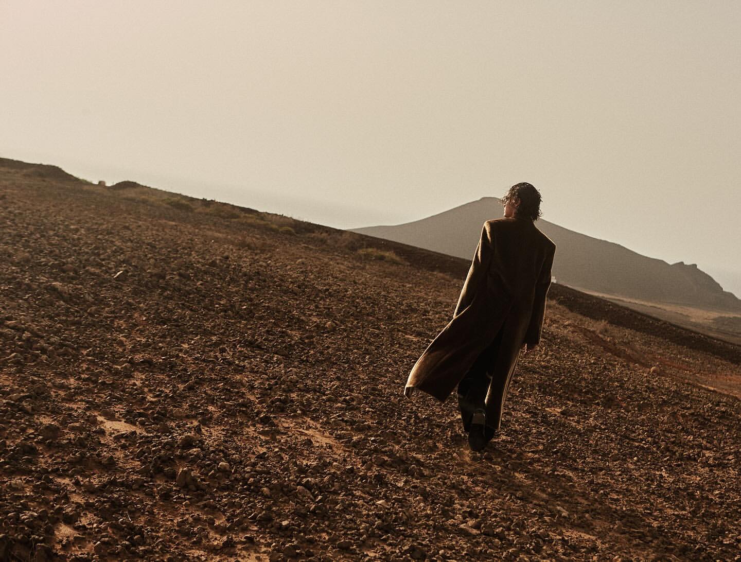 A person in a long coat walks across a rocky hillside, viewed from behind, with muted desert colors and mountain silhouettes.