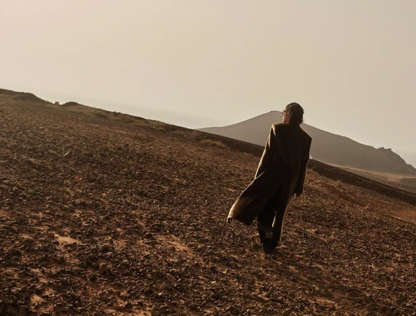 A person in a long coat walks across a rocky hillside, viewed from behind, with muted desert colors and mountain silhouettes.