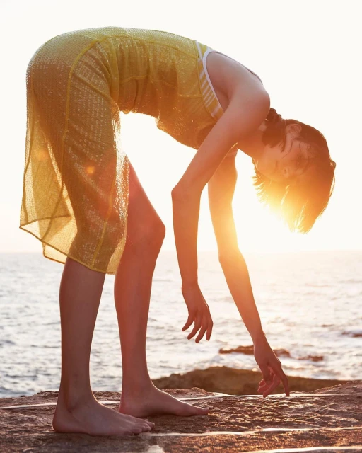 Young woman in a yellow dress bends forward at the edge of the ocean during sunset. The sun is glowing and the sky is bright.
