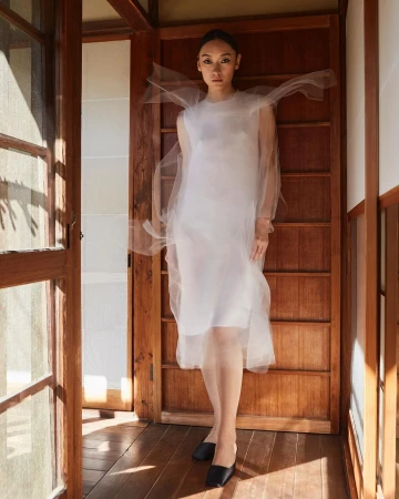 Woman poses in a white dress by a doorway with natural light casting shadows on the wooden floor in a fashion editorial shot.