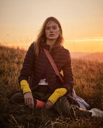 Model wearing a dark quilted puffer jacket and bright yellow wrist warmers sitting in a grassy meadow at sunset by Tassili Calatroni