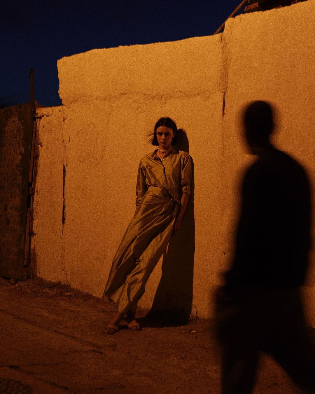 Woman in a stylish jumpsuit poses against a textured wall in an urban alleyway at night, illuminated by artificial light and casting a shadow.