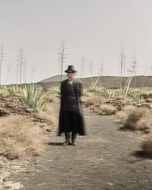 Person in dark clothing and hat stands on path in desert landscape with mountain, succulents, and dried shrubs under pale sky.