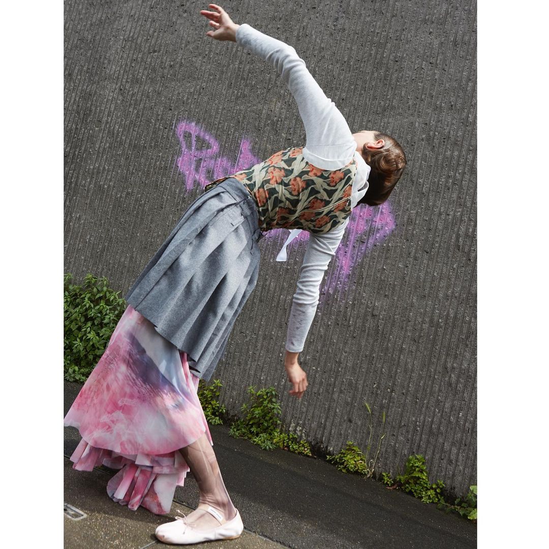 A woman in a printed top, grey skirt, and white shirt strikes a pose against a concrete wall outdoors, showcasing an editorial fashion look.