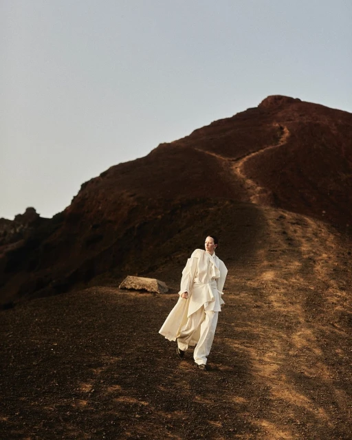 Adult woman in a white outfit walks down a brown dirt path on a hillside in natural light with a mountain in the background.