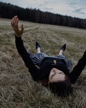 A person lying on dry grass in a field with arms raised, wearing a dark top and grey skirt against a distant treeline.