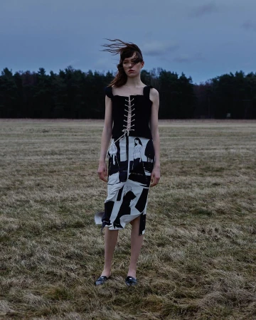 A model wearing a corset and patterned skirt stands in a field. Overcast sky and bare trees in the background. Fashion editorial.