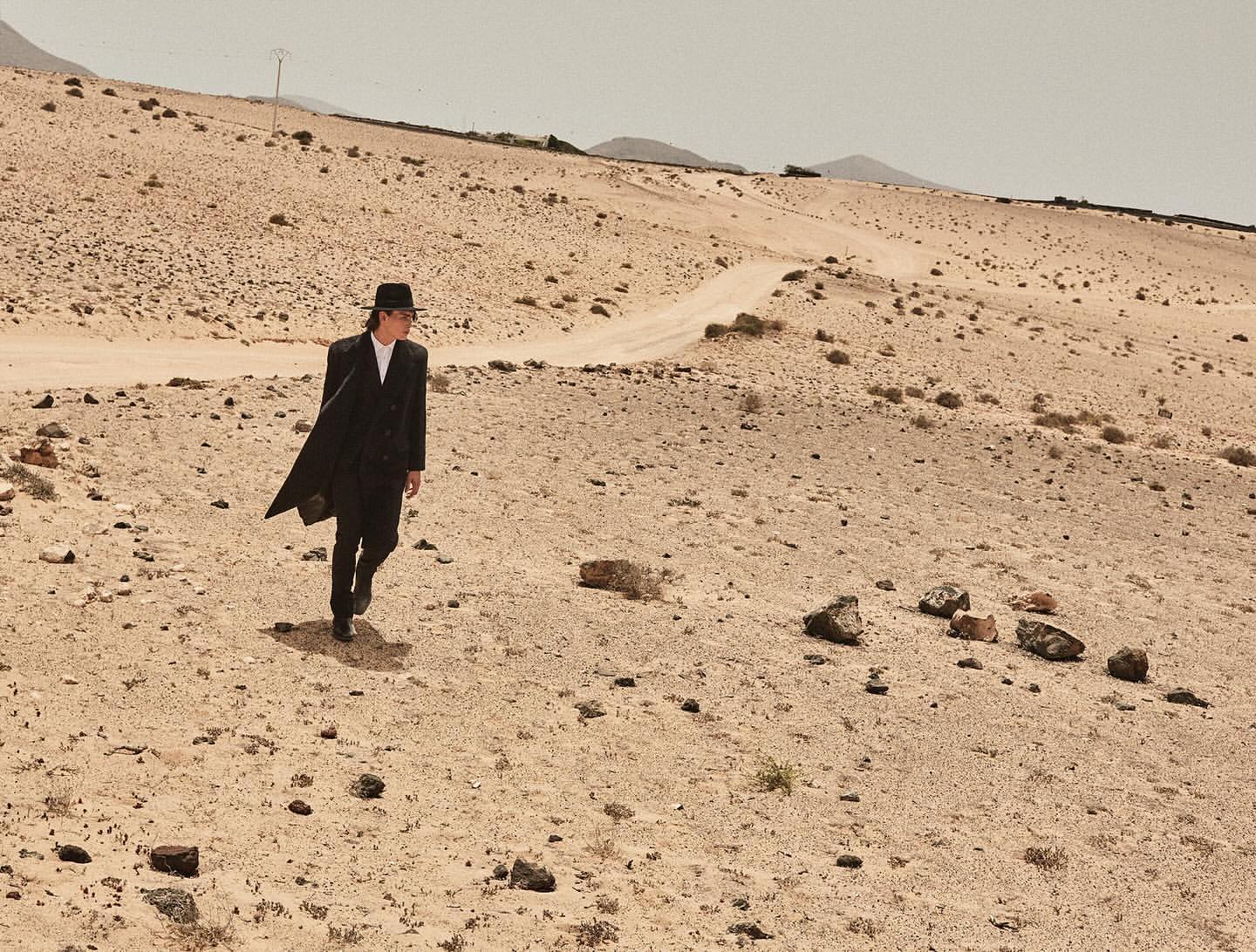 An adult male in a dark coat and hat walks across a desert landscape with mountains in the background and a pale sky overhead.