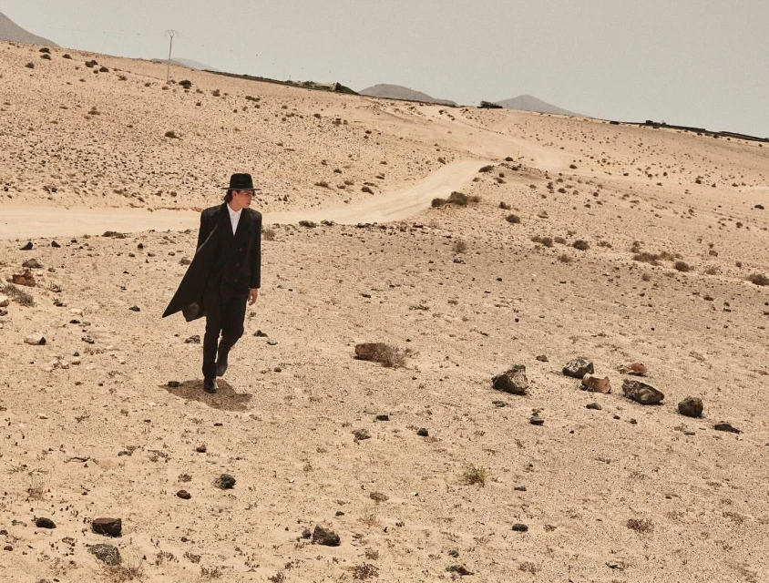 An adult male in a dark coat and hat walks across a desert landscape with mountains in the background and a pale sky overhead.
