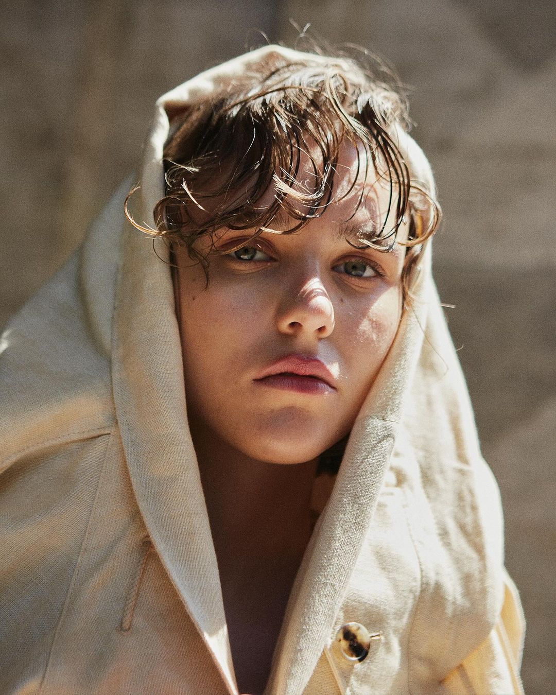 Close-up portrait of a person wearing a hooded cream coat with wet-look hair against a textured background by Tassili Calatroni.
