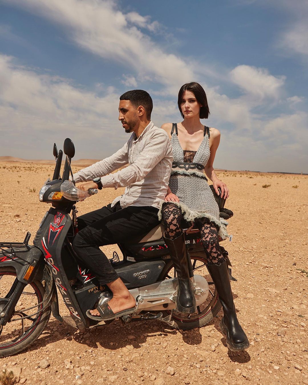 Editorial fashion shot featuring a male rider and female passenger on a scooter in a desert landscape under a bright sky with clouds.