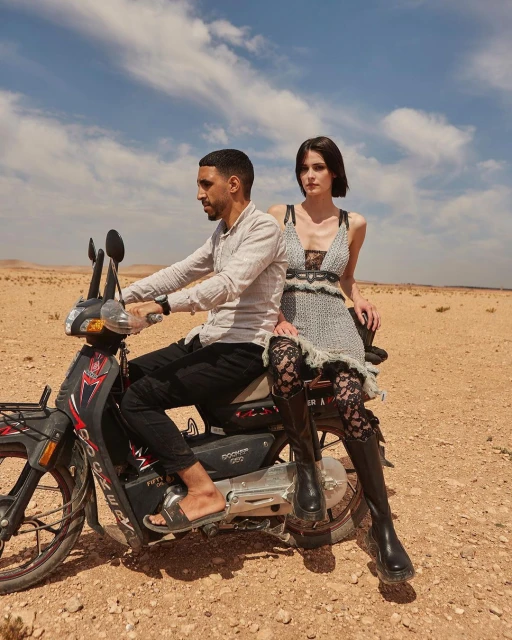 Editorial fashion shot featuring a male rider and female passenger on a scooter in a desert landscape under a bright sky with clouds.