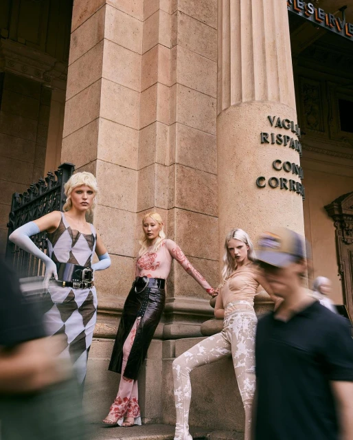 Three fashion models pose outdoors against a building with blurred pedestrians in the foreground, showcasing alternative couture and style.