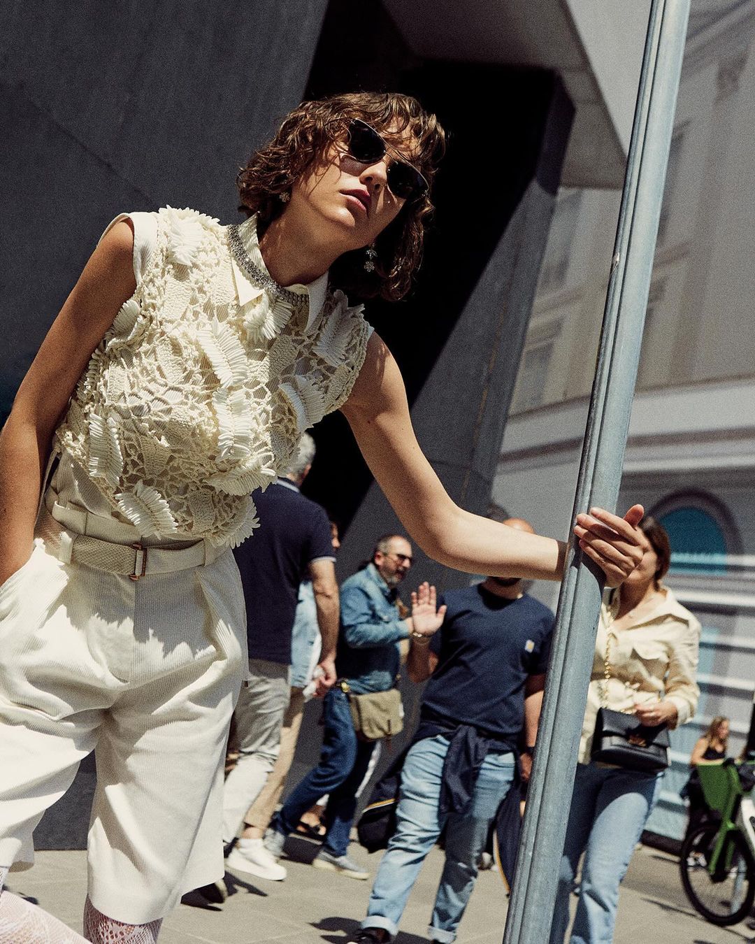 Model in cream lace top and white pleated shorts wearing sunglasses leaning on a pole on a busy street by Tassili Calatroni