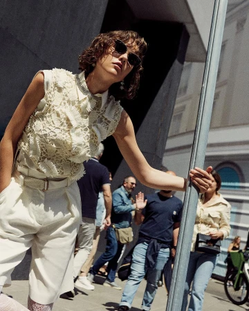 Model in cream lace top and white pleated shorts wearing sunglasses leaning on a pole on a busy street by Tassili Calatroni