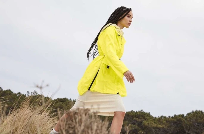 Woman in a bright yellow hooded jacket walking outdoors past dry coastal brush against an overcast sky.