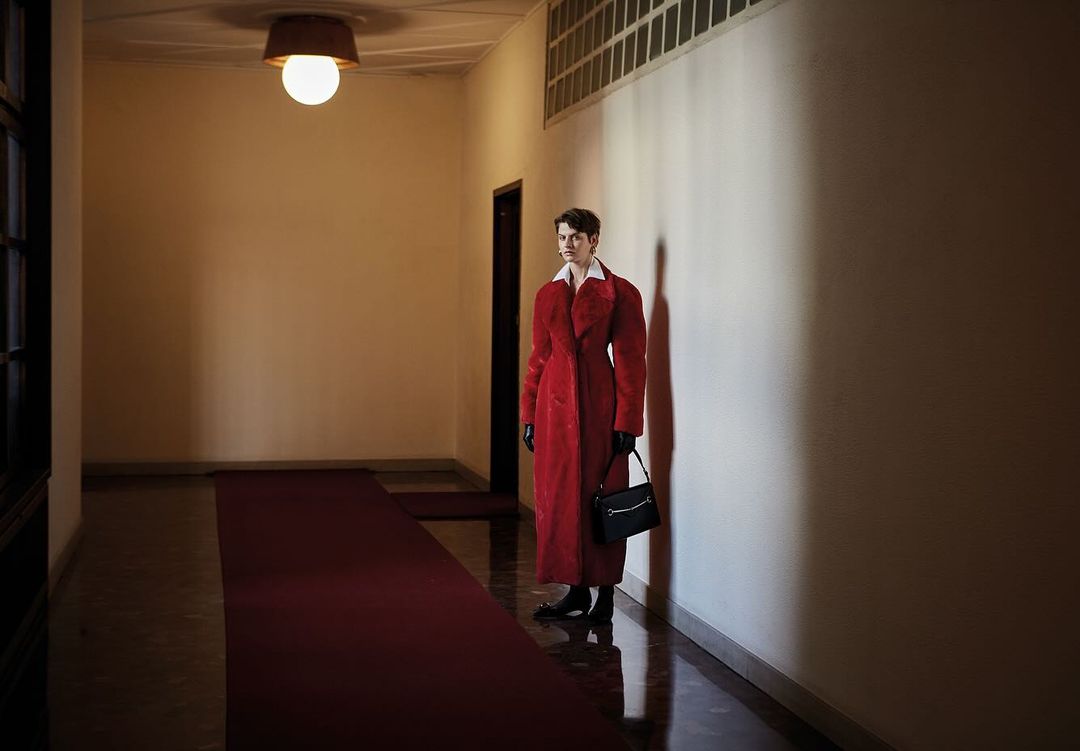 Woman in a red coat stands in a hallway with a carpet, holding a black purse, with warm lighting on the wall, and indoor setting.