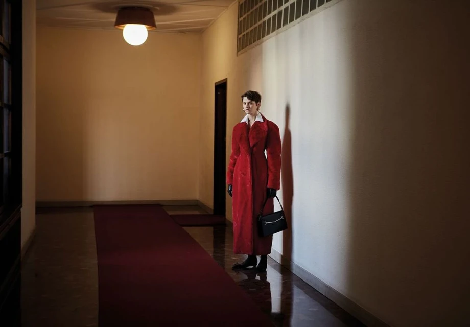 Woman in a red coat stands in a hallway with a carpet, holding a black purse, with warm lighting on the wall, and indoor setting.
