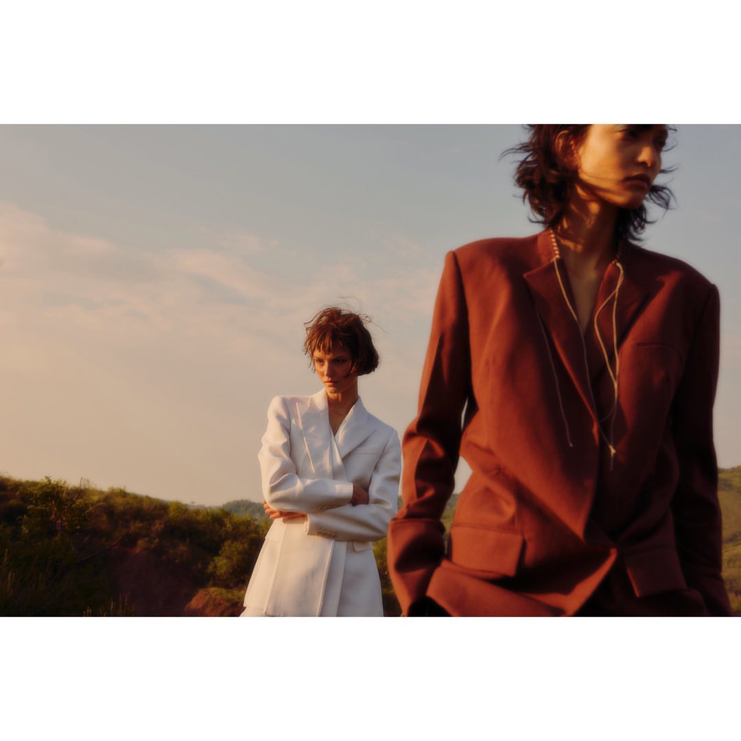 Two women in jackets pose in a sunny outdoor setting, one in white and one in rust, showcasing modern fashion against a natural backdrop.
