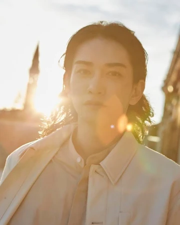 Close-up portrait of an adult with long hair in a cream-colored jacket, set against a sunlit urban backdrop with building silhouettes.