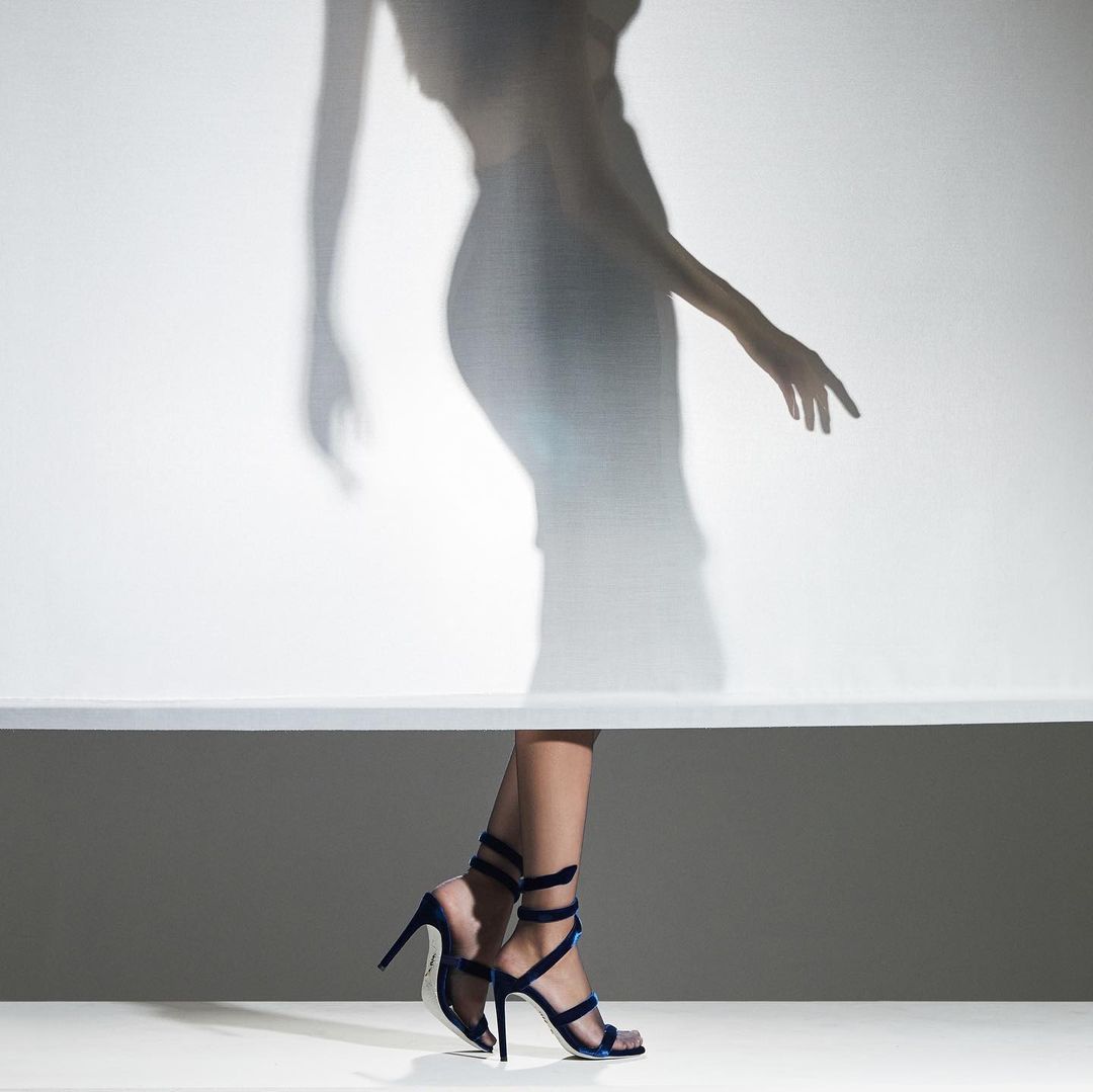 Fashion shot featuring woman's legs in blue stiletto heels against a graduated background with the blurry shadow of a figure above.