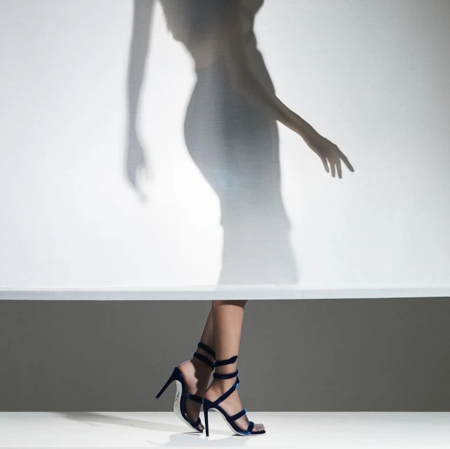 Fashion shot featuring woman's legs in blue stiletto heels against a graduated background with the blurry shadow of a figure above.
