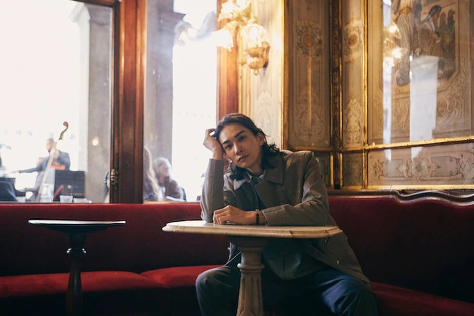Indoor portrait of an adult male with long dark hair resting at a marble table with a red sofa in an ornate, vintage-style restaurant.