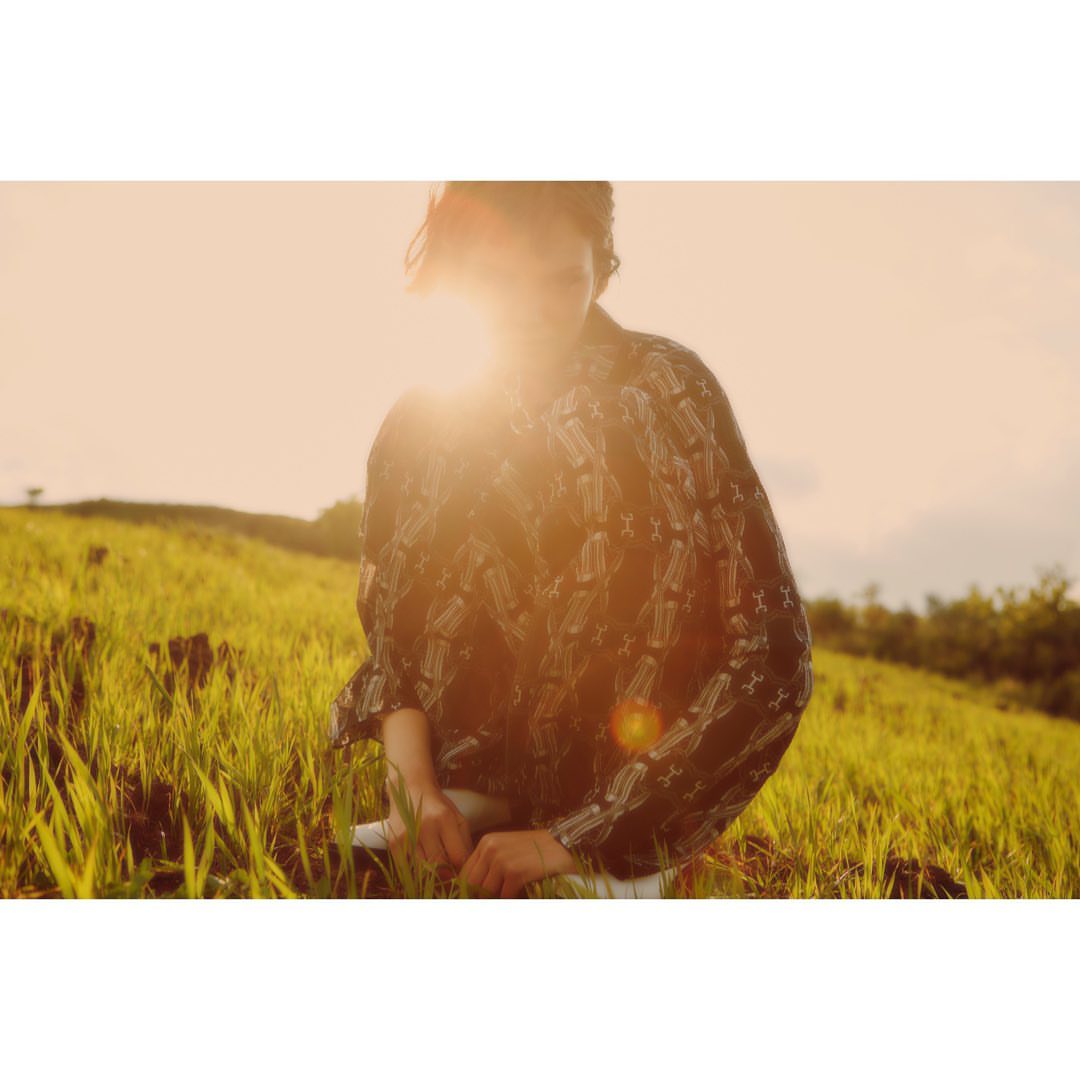 Woman in patterned shirt sits in a grassy field during golden hour, bathed in warm light with lens flare, serene outdoor setting.