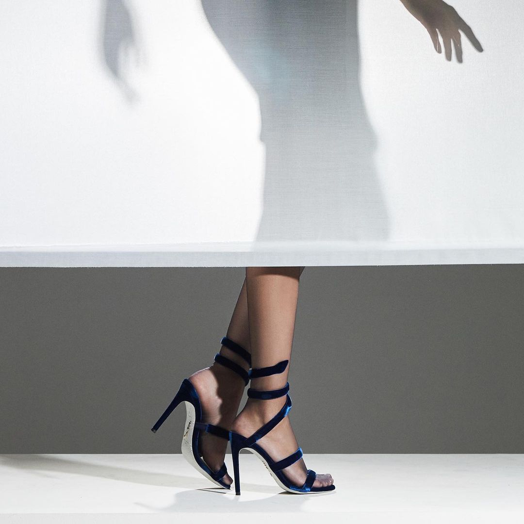 Woman's legs wearing blue velvet high heel sandals with a shadow figure against a white backdrop, showcasing fashion footwear.