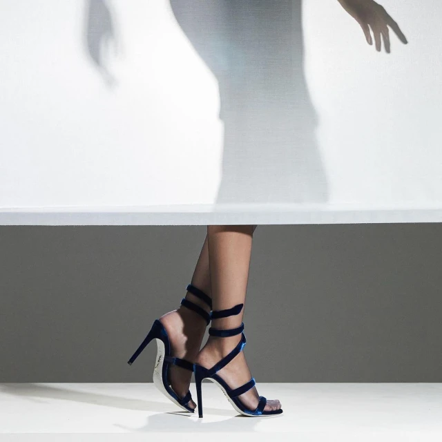 Woman's legs wearing blue velvet high heel sandals with a shadow figure against a white backdrop, showcasing fashion footwear.