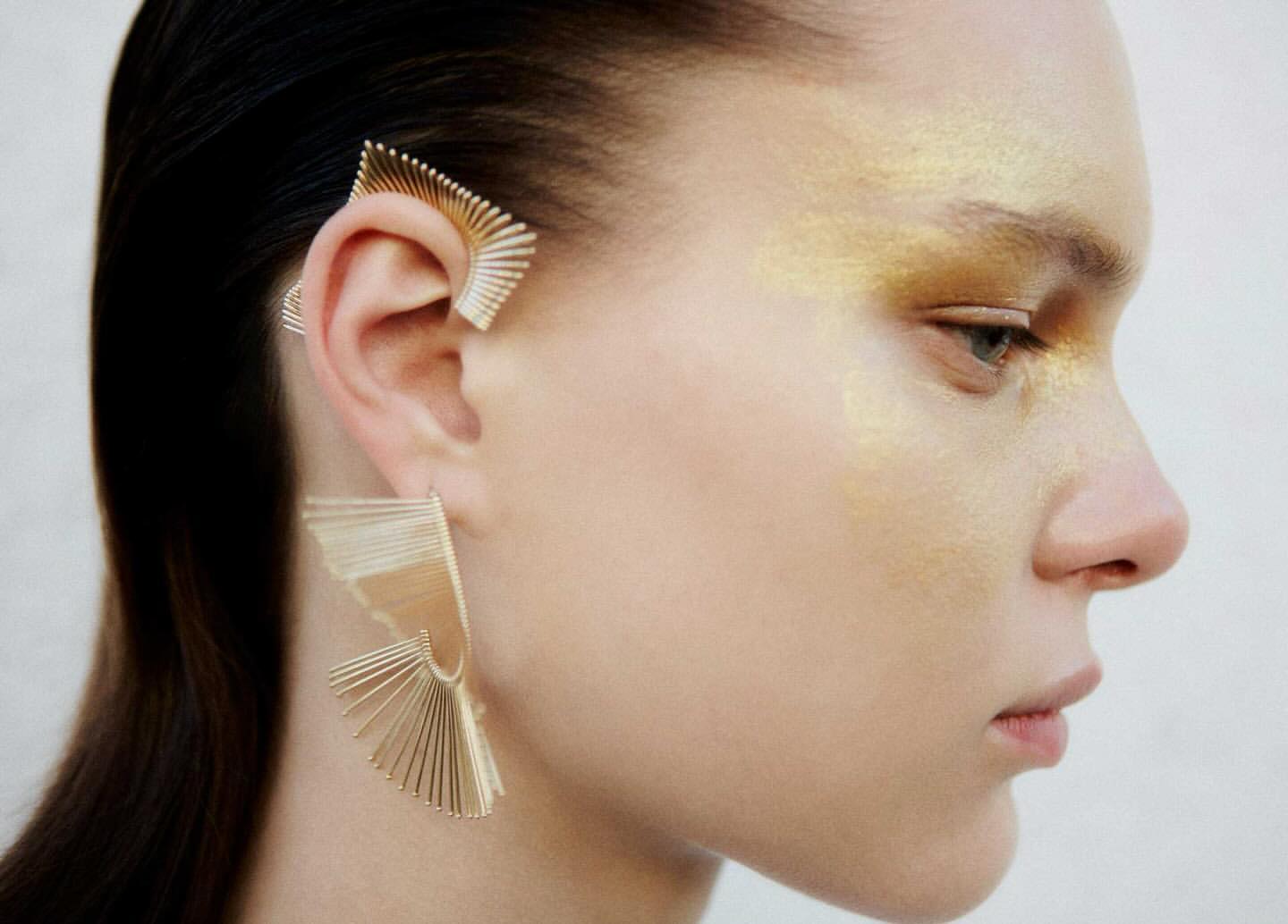 Close-up of a fair-skinned woman wearing artistic gold ear cuff and a matching earring, complemented by gold eyeshadow and dark hair.