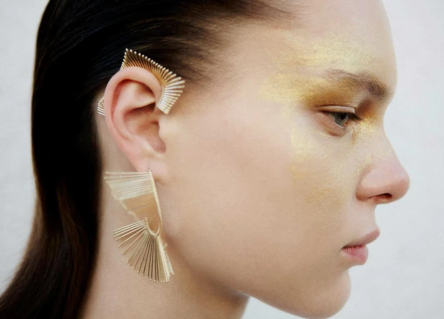 Close-up of a fair-skinned woman wearing artistic gold ear cuff and a matching earring, complemented by gold eyeshadow and dark hair.