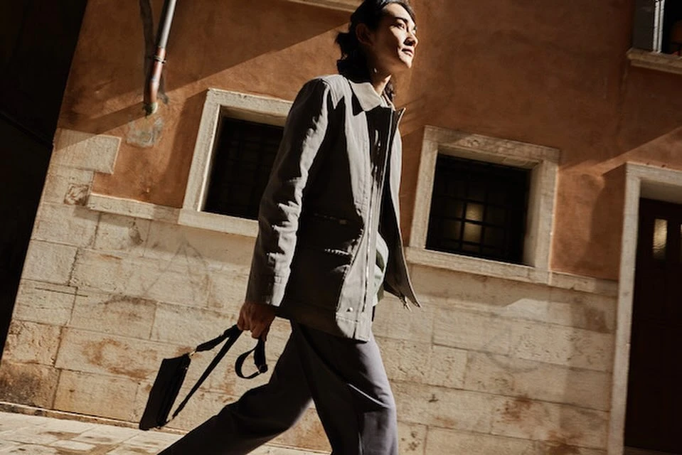 Adult male walking in a gray coat and pants while holding a briefcase in a city street with old stone architecture in the background.