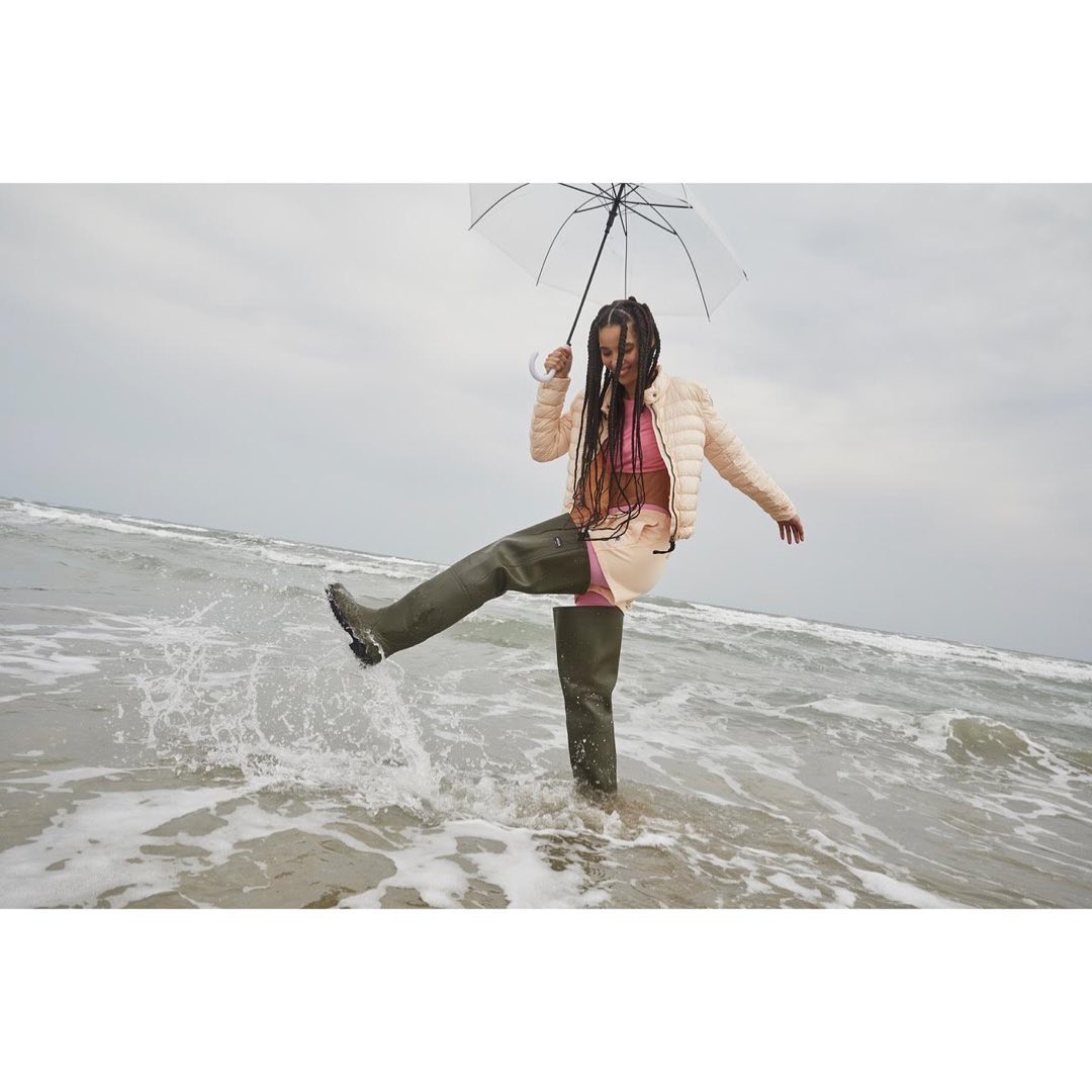 Woman in pink top and beige jacket kicking water in olive green boots with transparent umbrella on the beach during daytime.