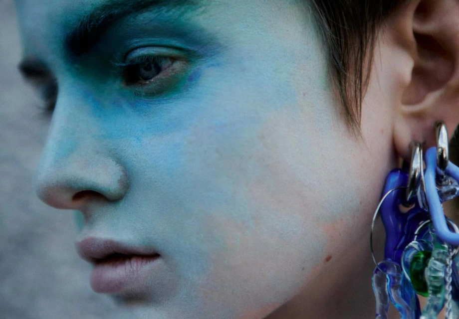 Close-up portrait of a person with blue face paint and multiple earrings, set against a blurred background. Beauty shot.