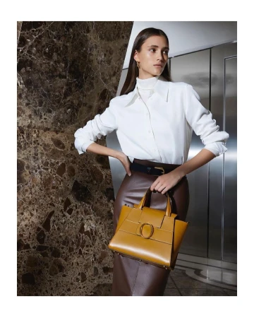 Model in white shirt and brown skirt holding a yellow leather handbag against marble and metal interior