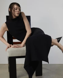 Woman with wet dark hair in a black dress seated on a wooden chair against a plain light wall