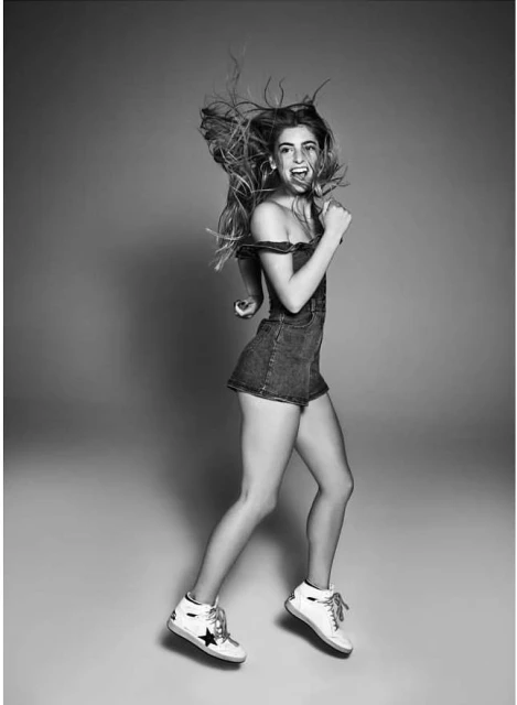 Full-length shot in black and white of a young woman jumping with joy in a studio wearing a denim romper and sneakers.