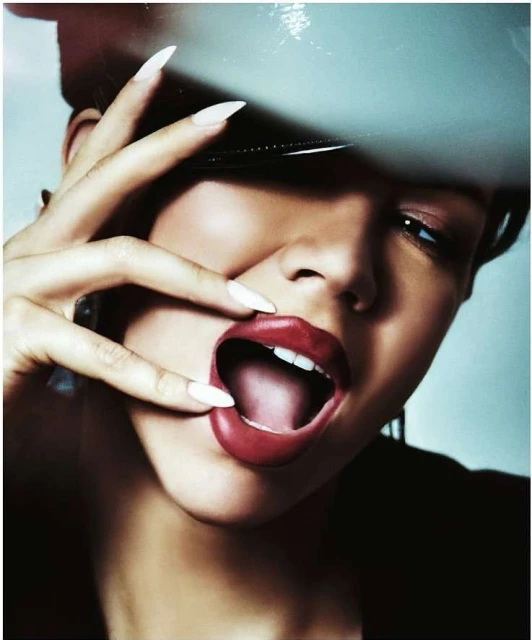 Close-up studio portrait of a woman with white nails and dark hair wearing a patent leather hat with a large mouth expression.