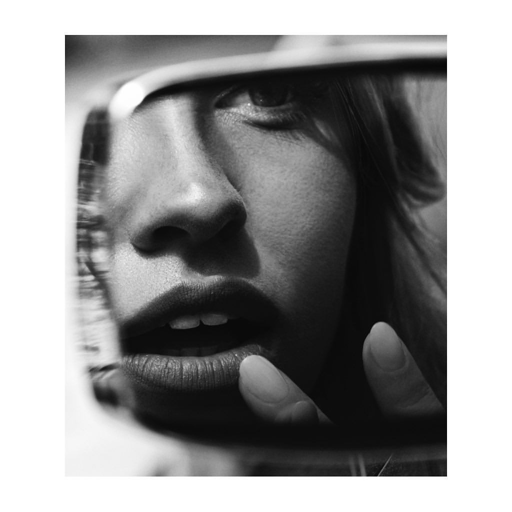 Close-up black and white photo of a woman's face reflected in a mirror, showing her eye, nose, and mouth with a finger touching her lip.