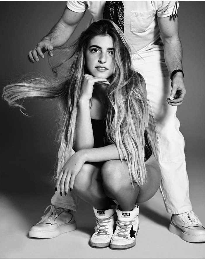 Black and white studio shot of a young woman with long hair, crouching, in front of a man with his hands near her hair.