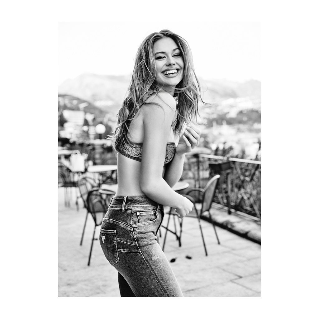 Smiling woman in blue jeans and a bra top standing on a terrace with a cityscape in the background, black and white portrait.