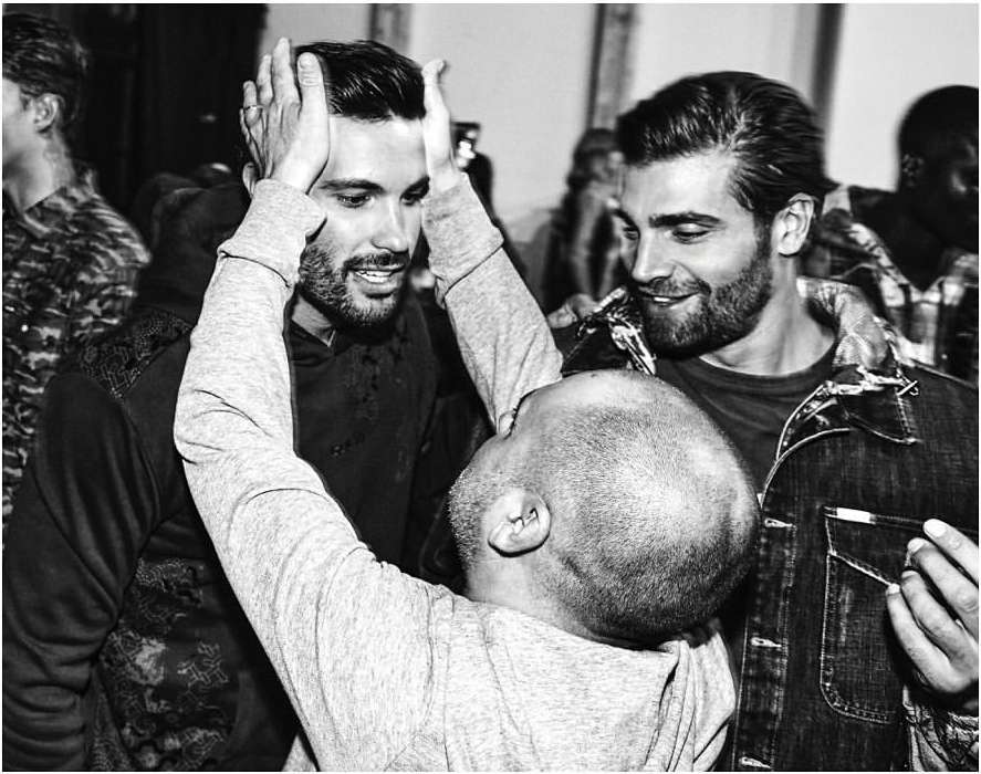 Black and white image of two adult men with beards smiling as a third person styles one man's hair, indoor setting, candid.