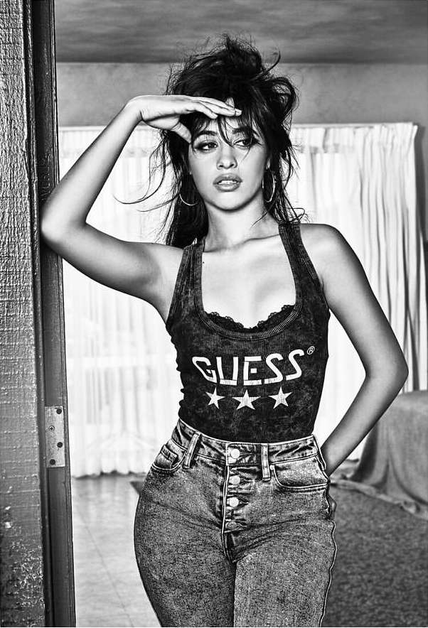 Black and white studio portrait of a woman in a Guess tank top and high-waisted jeans, gazing into the distance with her hand on her forehead.