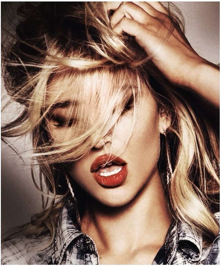 Close-up of a blonde woman with red lipstick and a plaid shirt, hand in her hair, creating a casual, stylish look with bright, bold makeup.