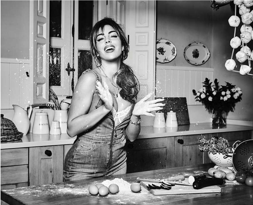 Black and white image of a woman in denim overalls clapping her hands in a kitchen covered in flour, surrounded by cooking ingredients.