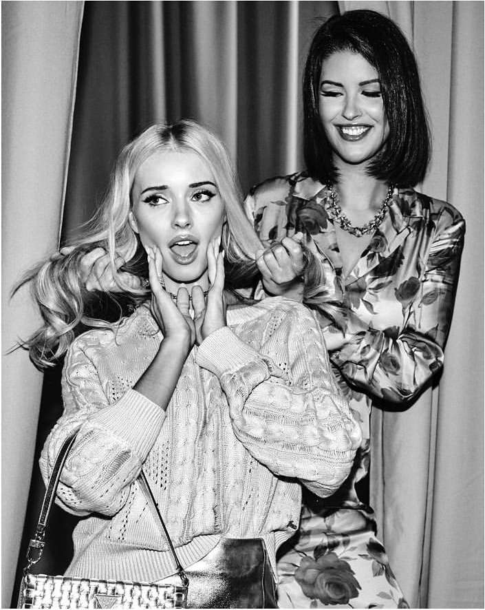 Two women pose indoors in a black-and-white photo, one with blonde hair and one with short black hair, wearing patterned tops.