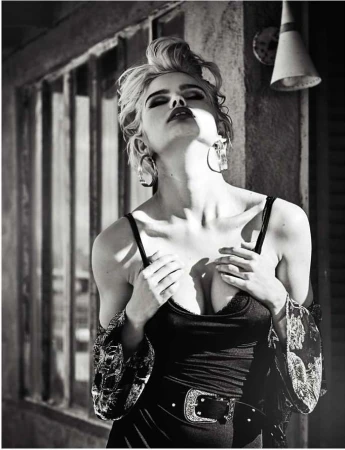 Black and white portrait of a woman with blond hair wearing a dark top, large hoop earrings, and a belt, tilting her head back outdoors.