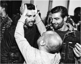 Candid black and white photograph of a man with his hands on another man's head, surrounded by smiling men in a gathering